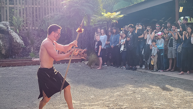 A photograph of a young man performing with a taiaha in traditional performance attire to an audience outside. 