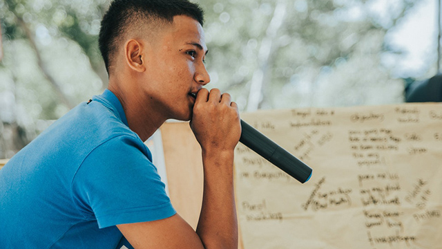 A photograph of a Māori boy holding a microphone to his mouth with a handwritten presentation behind him. 