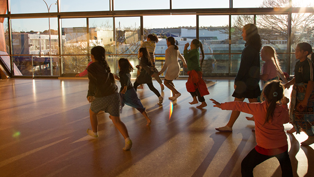 A photograph of a group of children running away from the camera into a sunlit dance studio. 