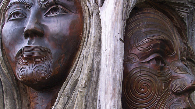 A photograph of driftwood carvings, showing two faces with facial tāmoko, a woman on the left and a man on the right. 