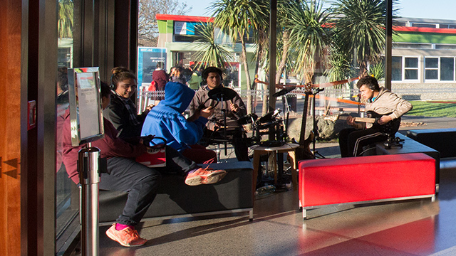 A photograph of five students sitting practising music, with the leftmost student playing a guitar, and another student playing a drumkit. 