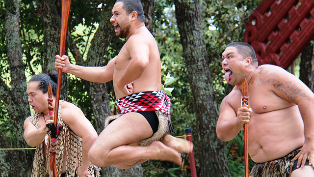 A photograph of three Māori men performing a haka. All performers are in traditional performance attire, and the middle performer is mid-air as he jumps. 