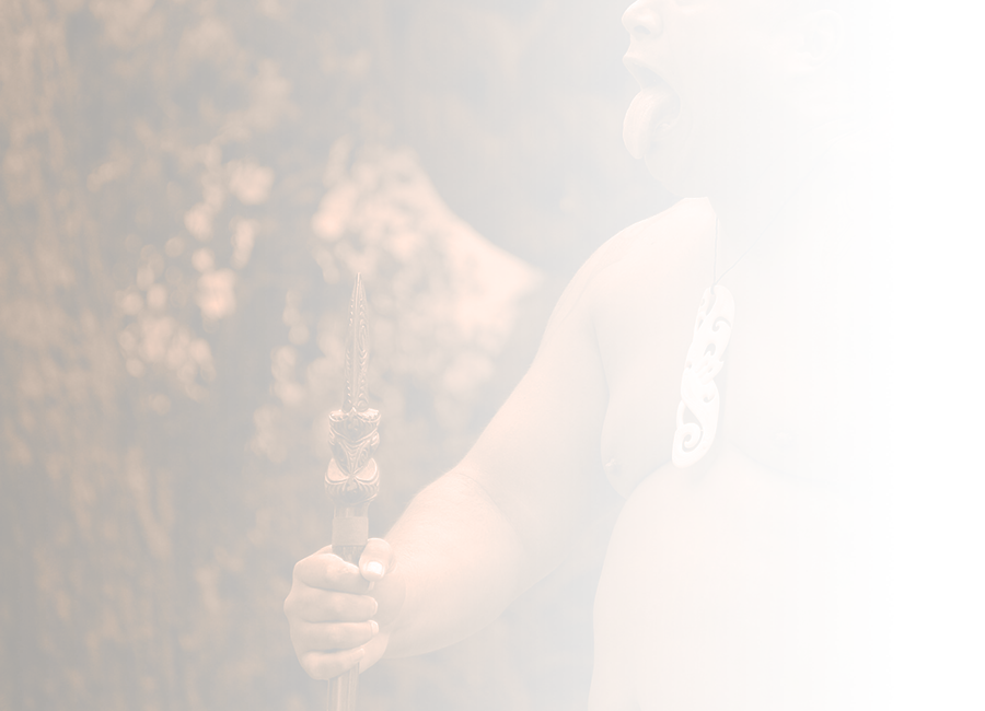 A photograph of a Māori man performing a pūkana, with a bone taonga, a taiaha to the left, and a tāmoko on his left arm. 