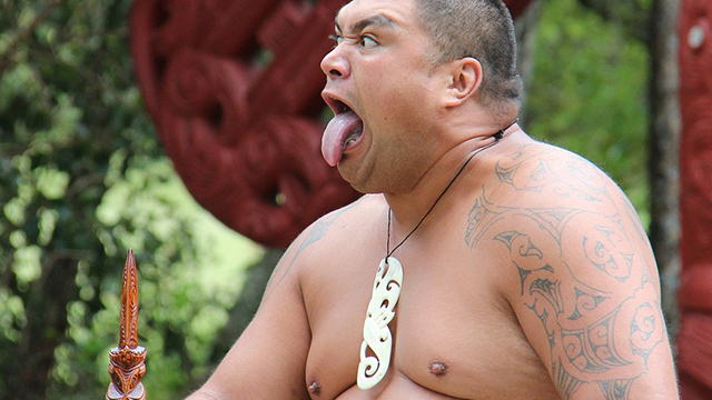 A photograph of a Māori man performing a pūkana, with a bone taonga, a taiaha to the left, and a tāmoko on his left arm. 