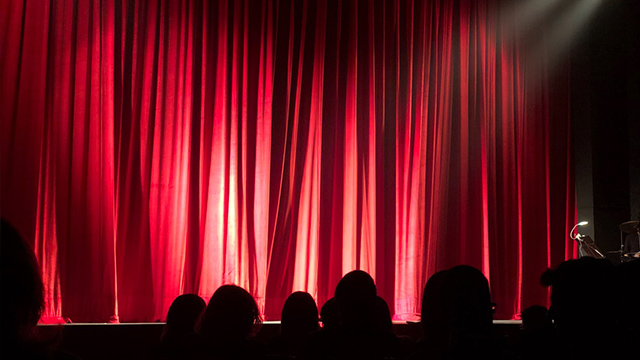 A photograph of the silhouette of an audience waiting in front of a stage obscured by red curtains. 
