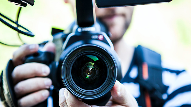 A photograph of a video camera facing the viewer directly, with the videographer out of focus behind the camera 