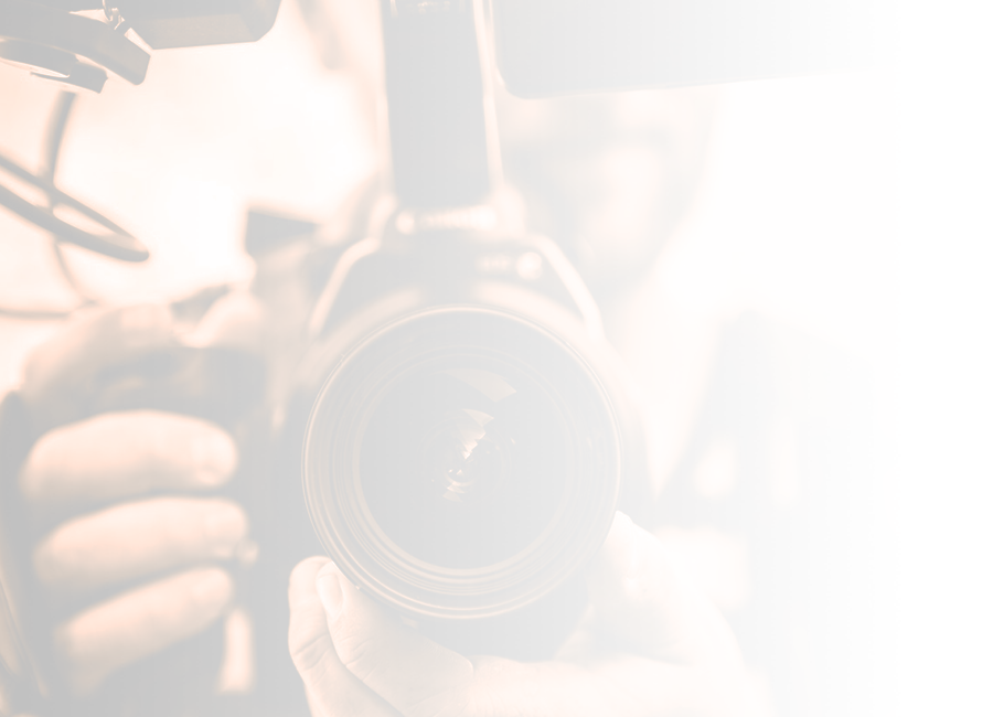 A photograph of a video camera facing the viewer directly, with the videographer out of focus behind the camera 