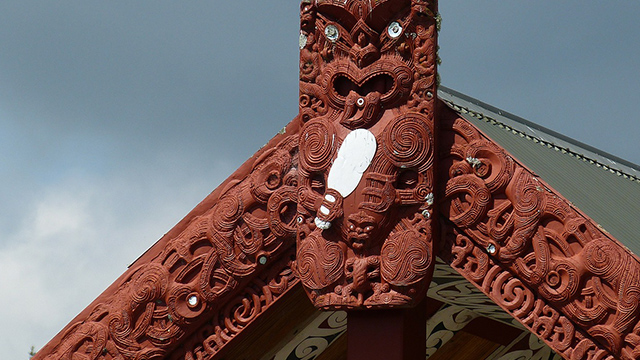 A cropped photograph of the top of a wharenui shows a tekoteko (carved figure), with the carved maihi (bargeboards) coming down from either side. 