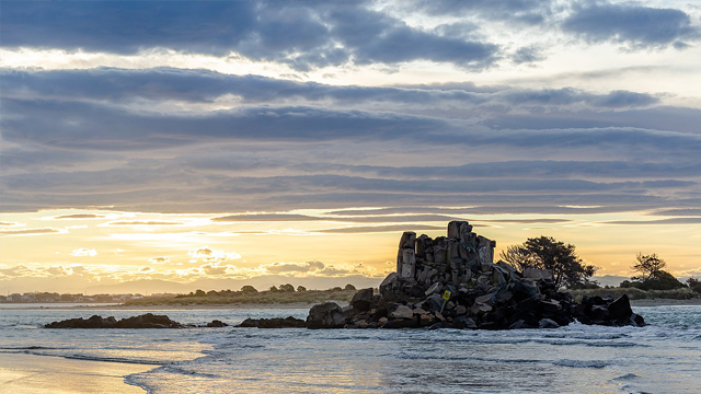 A beach at sunset. Boulders near the shore are silhouetted against a bright, cloudy sky